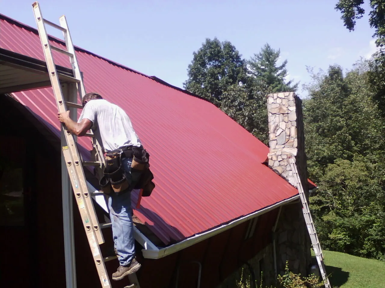 Roofer on a ladder beside a red metal roof and stone chimney — Soffit & Fascia Repair by Roofing Brothers in Mountain City